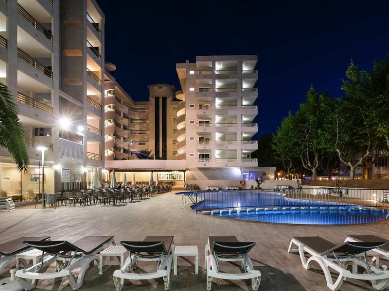 Night view of a lit hotel pool area with lounge chairs and surrounding buildings.