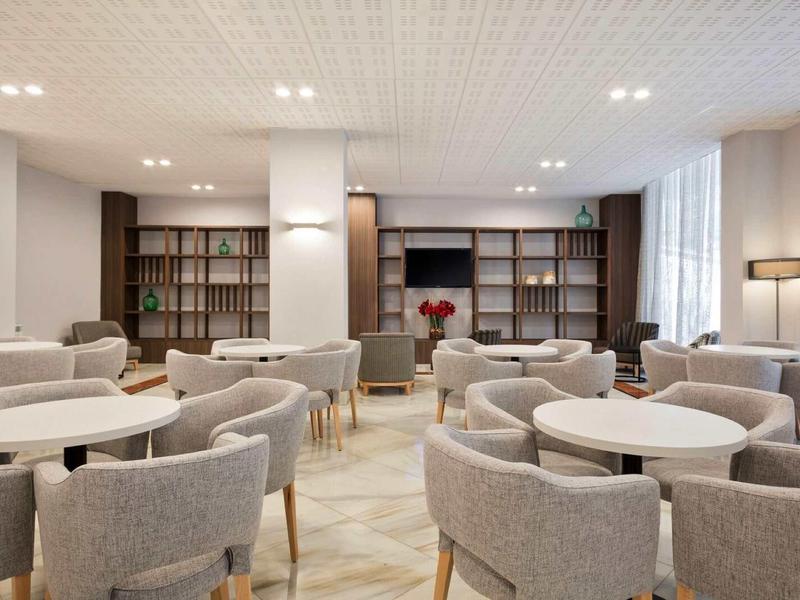 Modern hotel lounge with round tables, upholstered chairs, bookshelves, and natural light.