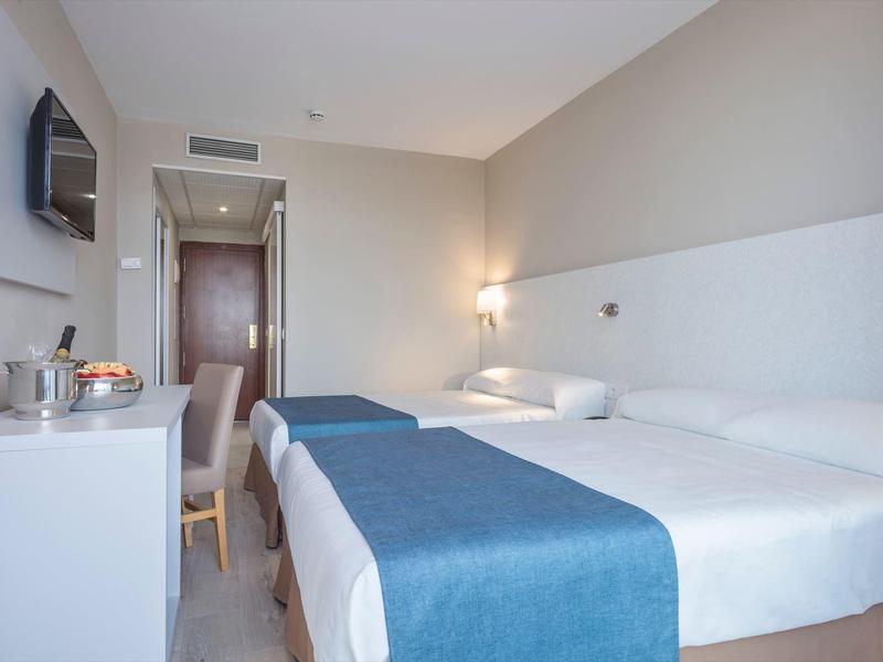 Světle zařízený hotelový pokoj se dvěma postelemi a modrými přehozy.