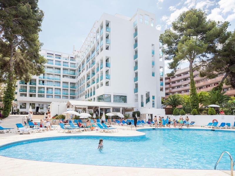 Moderní hotelový komplex s velkým venkovním bazénem obklopeným lehátky a vysokými stromy.