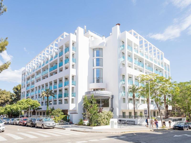 Moderní bílý hotel s velkými okny a balkony na slunném rohu ulice.