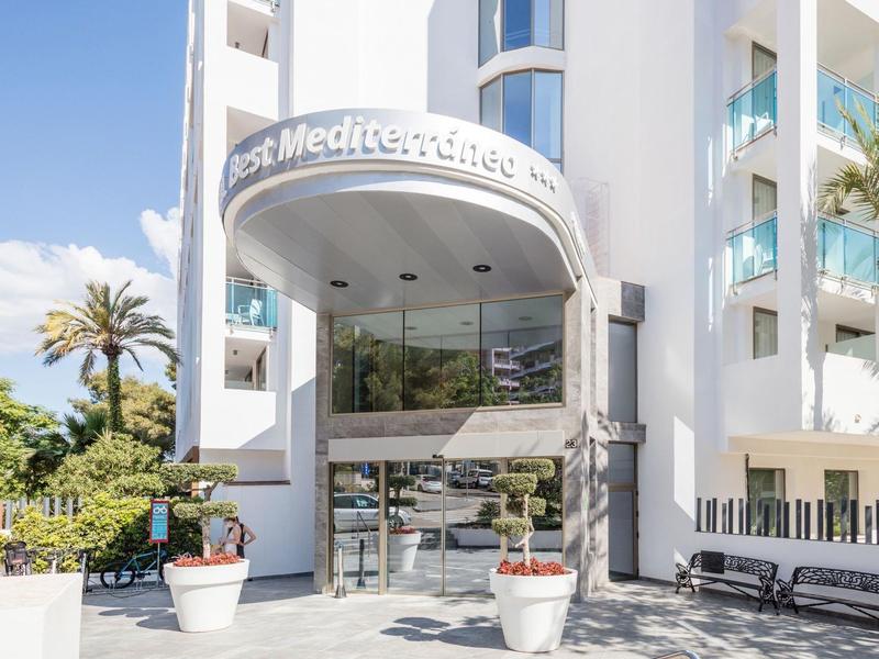 Vstup do moderního bílého hotelu s palmami a dvěma květináči.