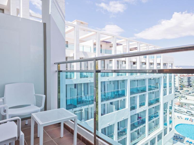 Balkon s bílými židlemi a skleněnou zábradlím s výhledem na moderní hotelovou budovu a bazén.