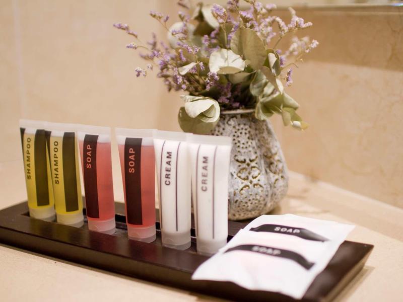 Sechs bunte Cremetuben und zwei weiße Tücher vor Vase mit getrockneten Blumen auf beige Ablage.