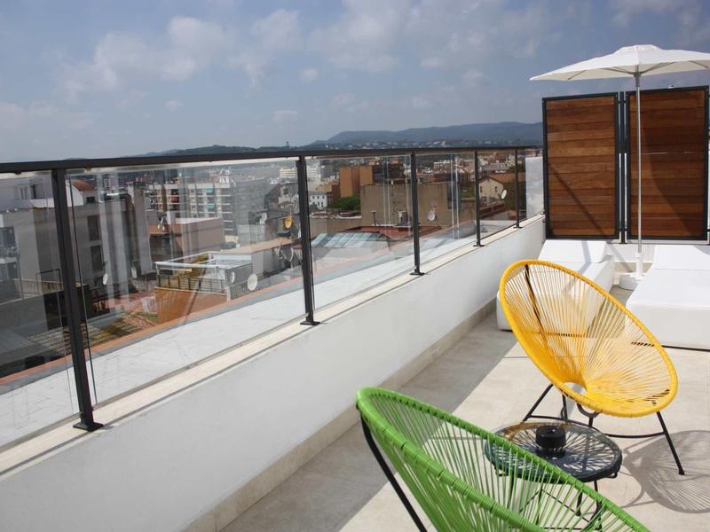Dachterrasse mit Glasgeländer, weißen Sitzmöbeln, bunten Stühlen und Sonnenschirm.