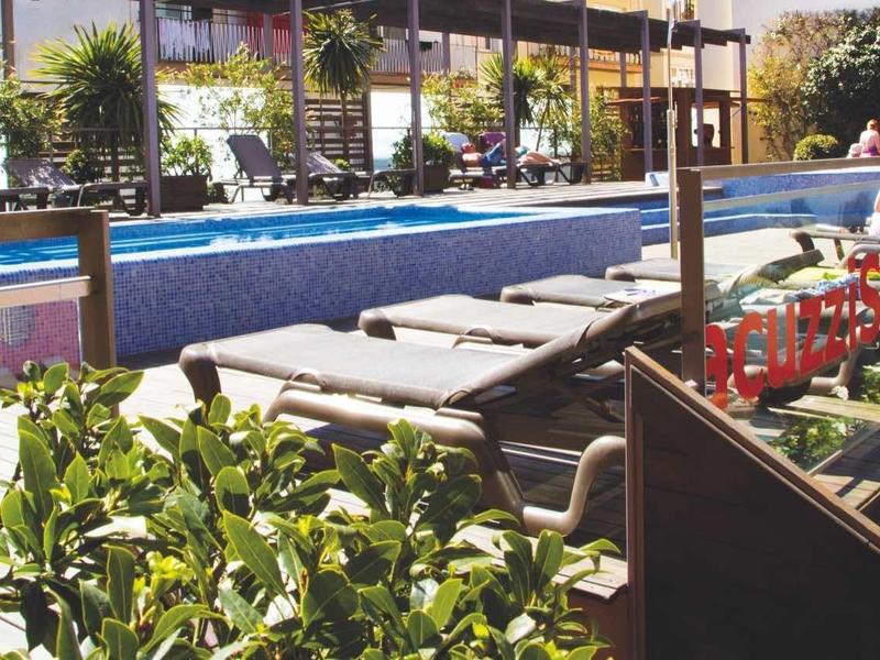 Außenpool mit blauer Wasserfarbe, Umrandung aus Holz, Liegen und grünen Pflanzen im Vordergrund.