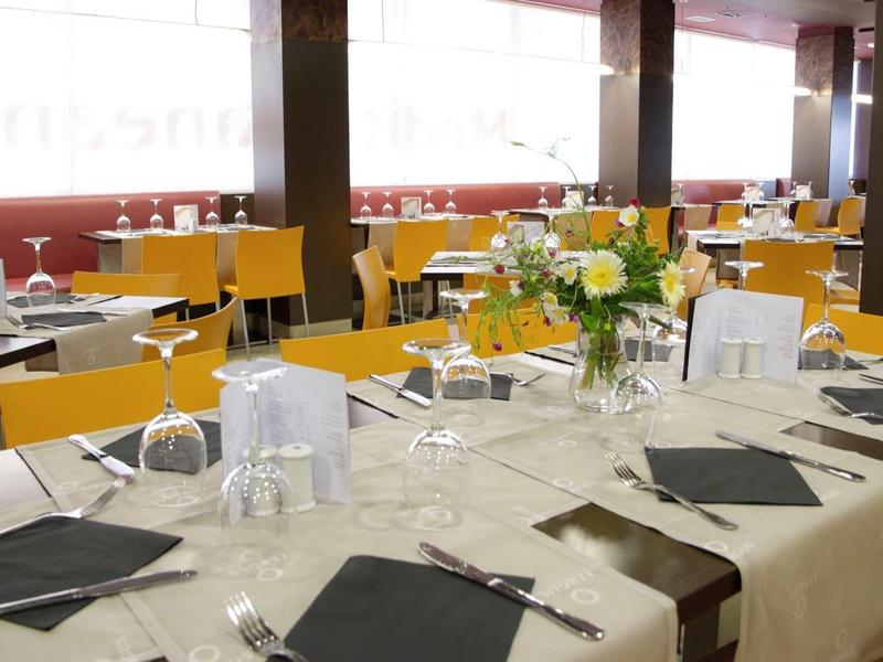 Großer Gastraum mit beige gedeckten Tischen, schwarzen Servietten und gelben Stühlen.