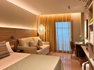 Modern hotel room with bed, sofa, desk, and warm lighting.