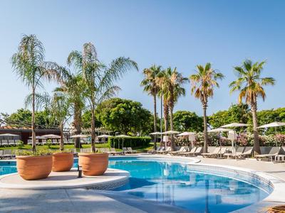 Round swimming pool with palm trees and sun umbrellas in a hotel garden under clear sky.