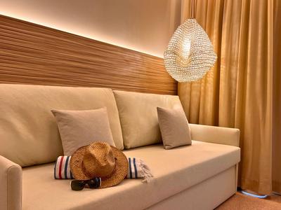Beige sofa with cushions, sun hat, sunglasses, and curtains in warm hotel room lighting.