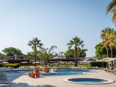 A well-maintained hotel pool area with umbrellas, palm trees, and seating under a clear sky.