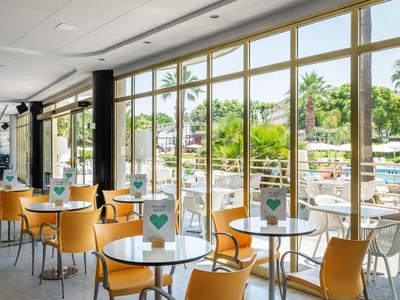 Modern indoor café area with white tables, orange chairs, and view of pool and palm trees.