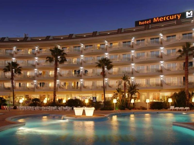 Hotelová budova s osvětleným bazénem a palmami v noci.