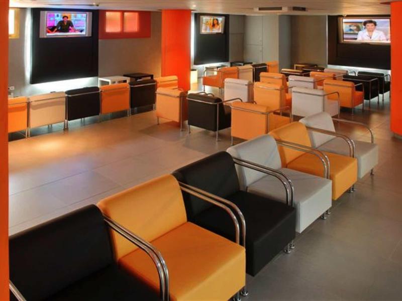Moderní čekárna s oranžovými, černými a bílými křesly a televizory na stěně.