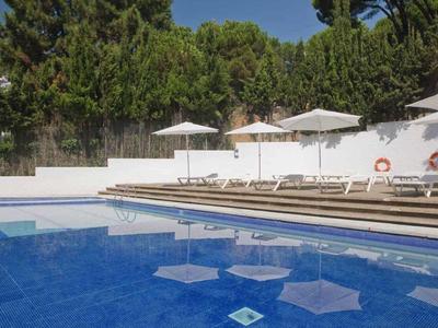 Bazén s modrou vodou, bílými lehátky a slunečníky vedle zdi a stromů.