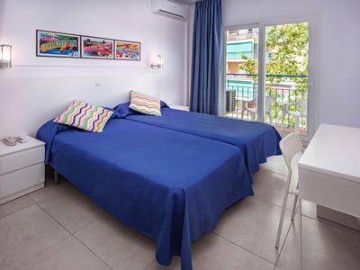 Světlo hotelový pokoj se dvěma postelemi, modrými povlečeními, balkonem a moderním nábytkem.