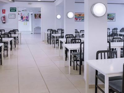 Světlá, moderní jídelna s bílými stoly a černými židlemi uspořádanými v řadách.