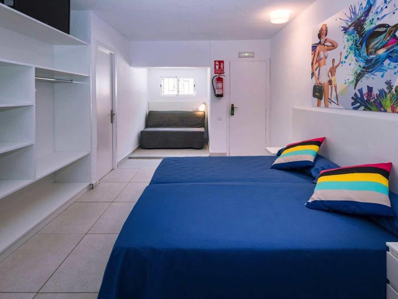 Moderní ložnice se dvěma modrými postelemi a barevnými polštáři, bílá stěna a police.