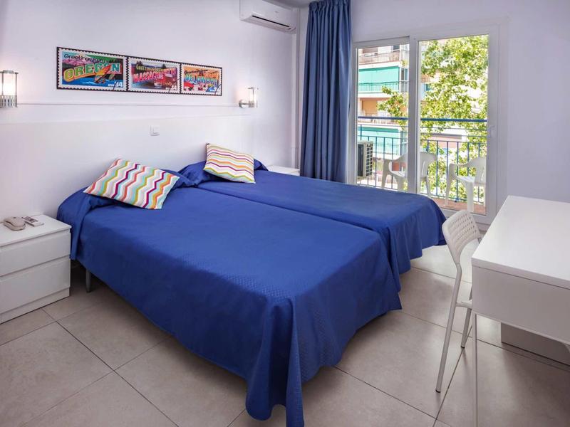 Světlo hotelový pokoj se dvěma postelemi, modrými povlečeními, balkonem a moderním nábytkem.