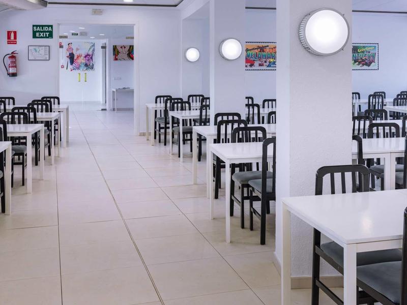 Světlá, moderní jídelna s bílými stoly a černými židlemi uspořádanými v řadách.