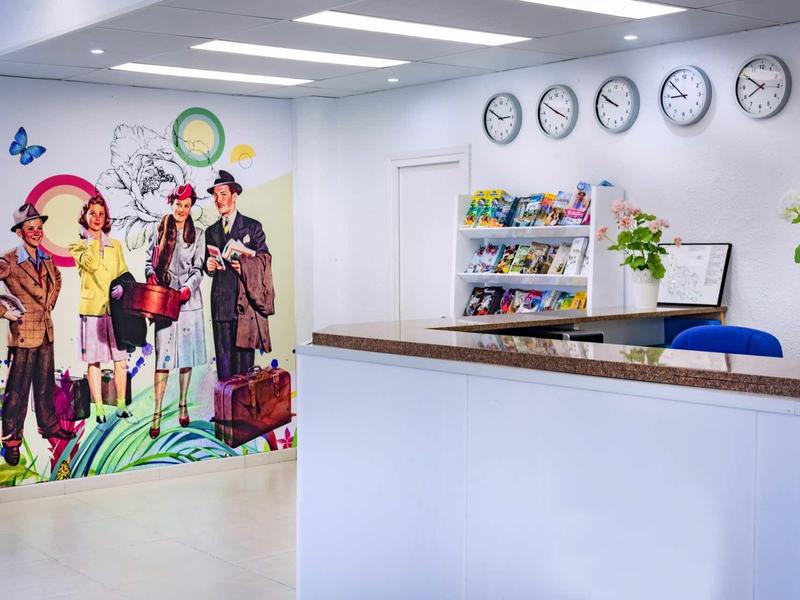 Světlá hotelová lobby s recepcí, barevnou nástěnnou malbou a několika hodinami.