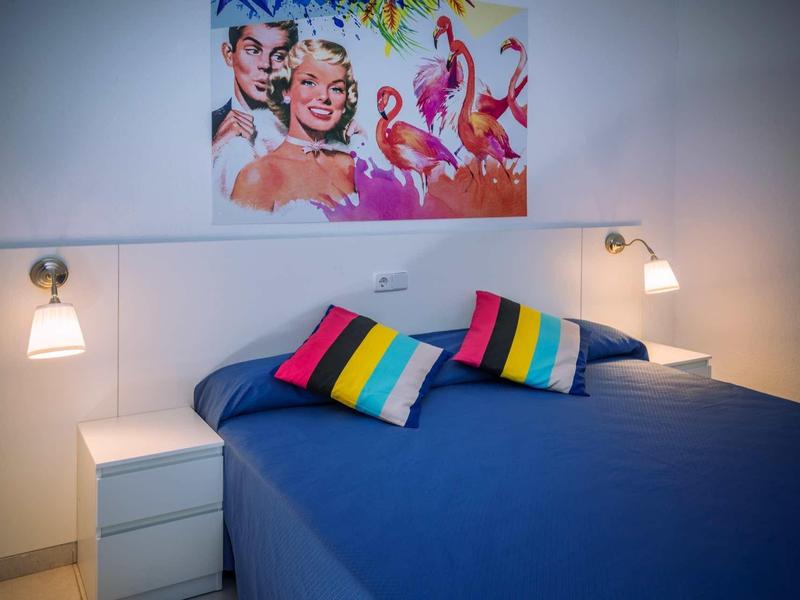 Ložnice s modrou postelí, barevnými polštáři, dvěma nočními stolky a obrazem s plameňáky.