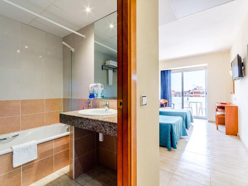 Helles Hotelzimmer mit Bad, gefliestem Boden, weiß-blauer Bettwäsche und Meerblick vom Balkon.