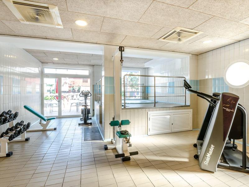 Heller Fitnessraum mit Geräten, Hanteln und großen Fenstern, weiß gekachelter Boden und Wände.