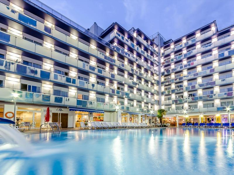 Großes beleuchtetes Hotelgebäude mit mehreren Etagen und hellem Pool im Vordergrund bei Abenddämmerung.