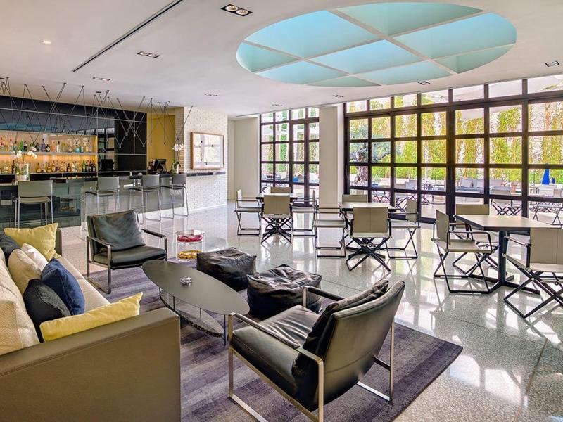Bright, modern hotel lounge with sofas, armchairs, bar, and large windows overlooking the garden.