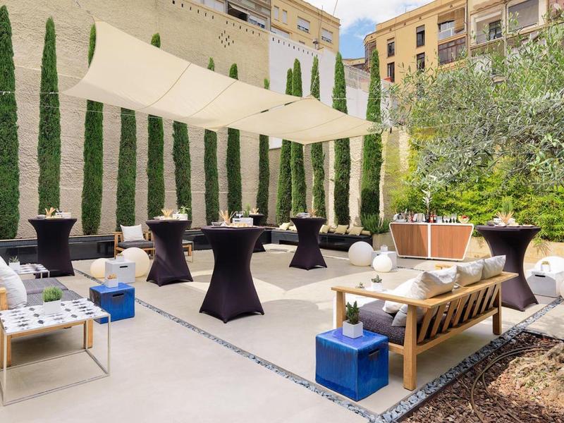 Modern outdoor area with seating and sun sails by a tall wall with cypress trees.