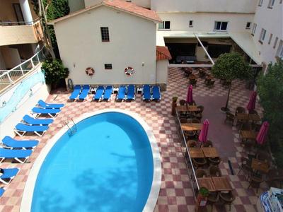 Widok na owalny basen hotelowy z niebieskimi leżakami i wyłożonym płytkami obszarem z stolikami i roślinami.