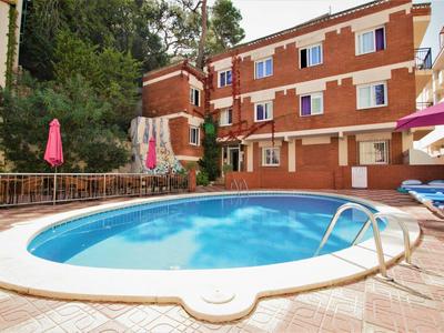 Okrągły basen na zewnątrz z czerwonymi parasolami i wielopiętrowym budynkiem hotelowym w tle.