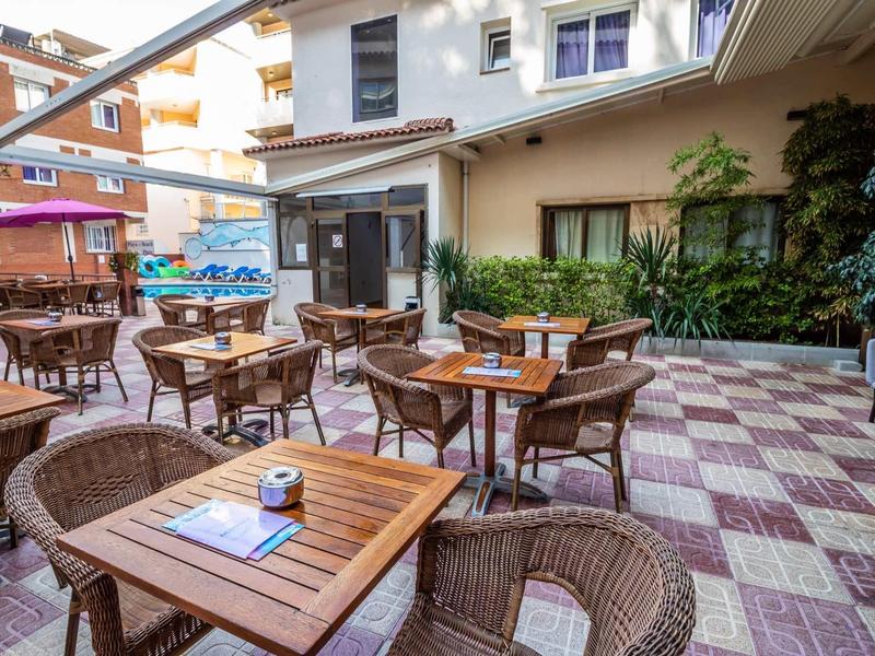 Dziedziniec hotelowy z drewnianymi stołami i krzesłami, otoczony roślinami i budynkami.