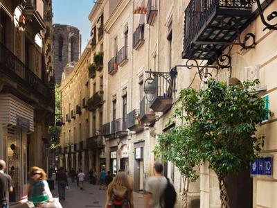 Enger, sonniger Stadtweg mit Passanten zwischen alten Gebäuden mit Balkonen und Pflanzen.