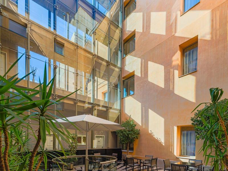 Cour ensoleillée avec murs en verre, plantes et sièges extérieurs avec tables et parasols.