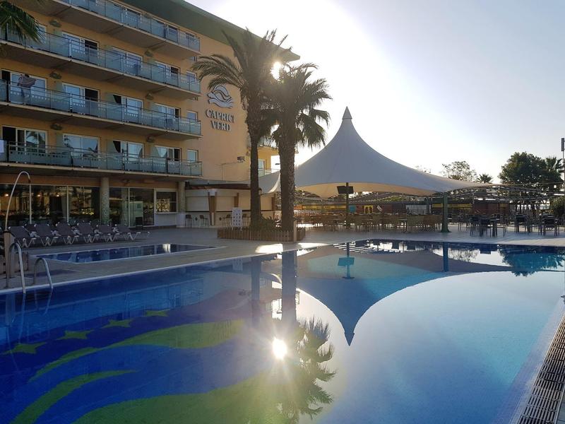 Area piscina dell'hotel con palme, tendalino e cielo azzurro alla luce del tardo pomeriggio.