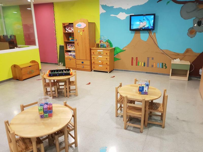 Sala giochi luminosa per bambini con tavoli in legno, sedie, scaffali e TV su una parete colorata.