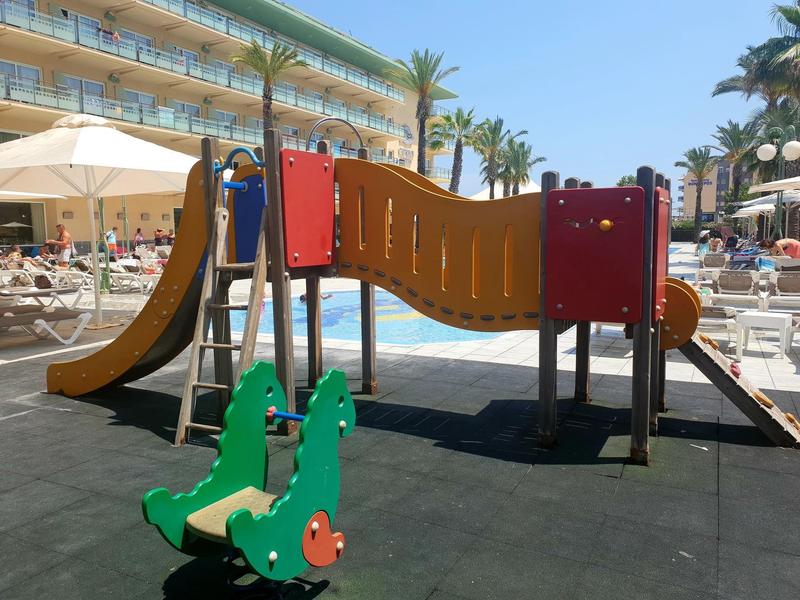 Area giochi per bambini con scivoli vicino a una piscina dell'hotel circondata da palme e lettini.