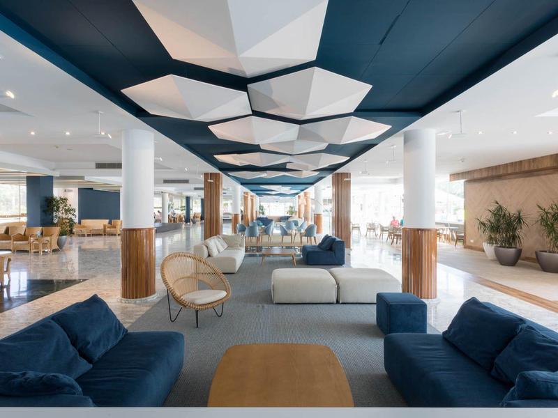 Moderna hall dell'hotel con divani blu, sedie in legno, soffitto geometrico e luce naturale.