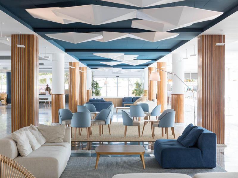 Hall di hotel moderno con sedute blu e beige, colonne in legno e soffitto geometrico.