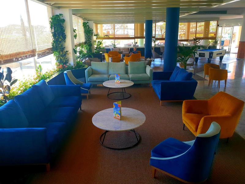 Lounge luminosa con divani blu e poltrone arancioni vicino a grandi finestre e piante.