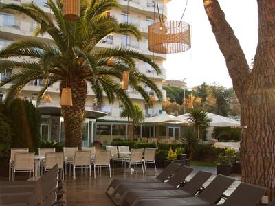 Terrasse d'hôtel avec chaises longues, tables et palmiers devant un bâtiment à plusieurs étages au coucher du soleil.