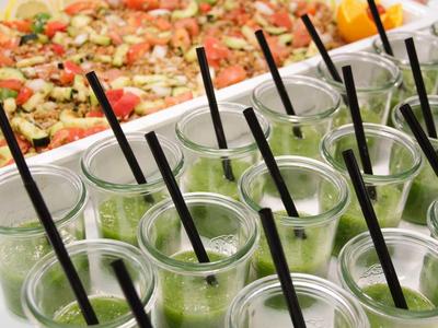 Plusieurs petits verres avec smoothie vert et pailles noires devant de grands plats de salade.