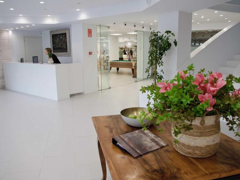 Hall d'hôtel lumineux avec réception moderne, escalier et arrangement floral décoratif sur table en bois.