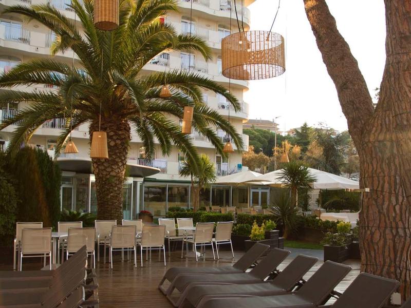Terrasse d'hôtel avec chaises longues, tables et palmiers devant un bâtiment à plusieurs étages au coucher du soleil.