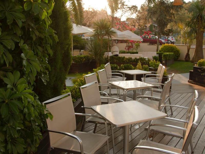 Terrasse avec tables et chaises blanches entourées de plantes vertes et de palmiers au soleil.