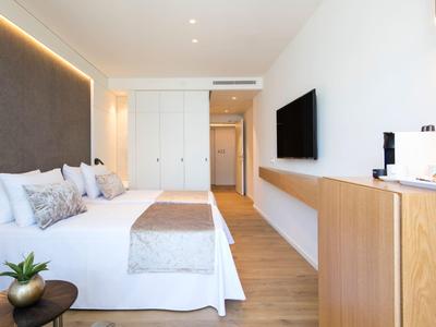 Nowoczesny pokój hotelowy z podwójnym łóżkiem, telewizorem i drewnianymi meblami w jasnym otoczeniu.