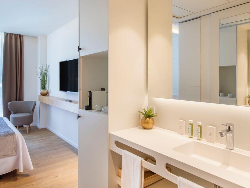 Nowoczesny pokój hotelowy z jasnymi ścianami, łóżkiem, biurkiem i umywalką z lustrem.