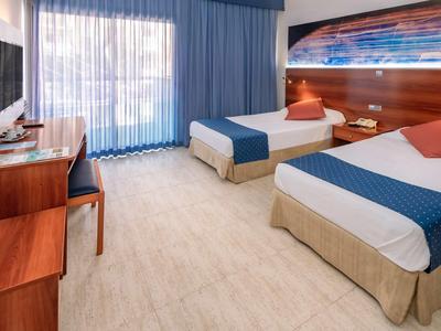 Pokój hotelowy z dwoma łóżkami, biurkiem, krzesłem i dużym oknem z niebieskimi zasłonami.